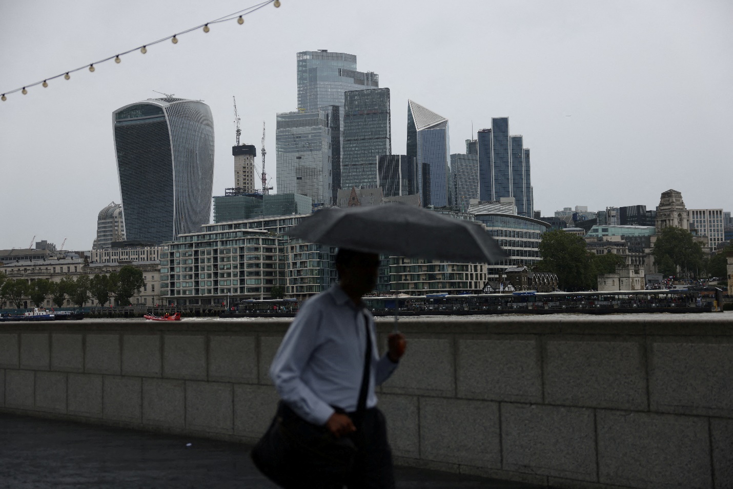 A pedestrian carrying an umbrella walks along the River Thames in view of City of London skyline in London