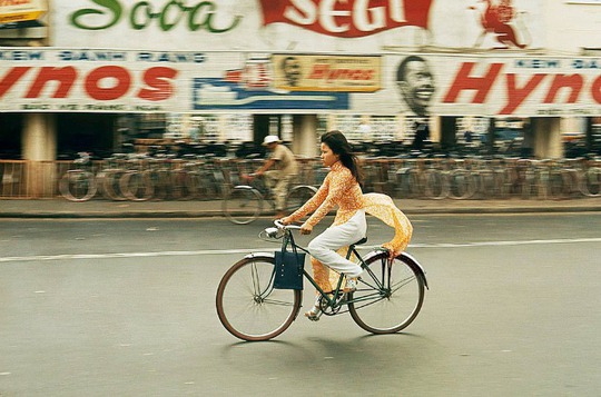 Một cô gái buộc tà áo dài vào chỗ ngồi sau xe để tránh vướng khi chạy xe đạp. Dọc hai bên đường lúc này là bảng hiệu quảng cáo của thương hiệu kem đánh răng phổ biến thời bấy giờ.